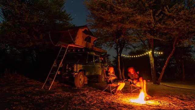 Static timelapse of adventurous couple on Africa Safari in Botswana Game Reserve, wild camping/glamping under moonlight night sky sitting around bonfire/fire, with truck, tent and solar lights.