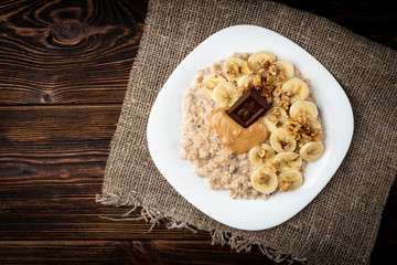 Oatmeal with banana, walnut, peanut butter and chocolate on dark wooden background.