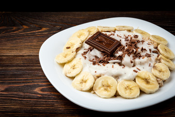 Oatmeal with banana, chocolate and yogurt on dark wooden background.
