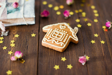 House shape ginger cookie with white glaze on brown wooden background. Golden star decoration and dry roses near cookie.