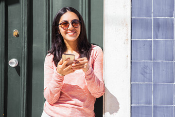 Happy woman using smartphone outdoors. Pretty young lady looking at camera and standing at door and wall. Communication concept. Front view.