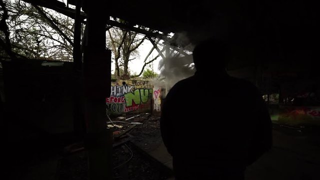 A young sillouetted millennial man standing in a dark abondoned building looking at graffiti and blowing smoke frome a vape machine, slow motion