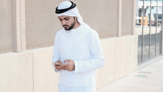Arab man using smart phone as he walks. Arabic uses mobile app  web browsing or chatting mock up while walking. Middle Eastern on Kandura dish dasha traditional clothing in the Gulf