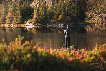 Obraz premium Fishing as recreation and sports displayed by fisherman at lake. Fishing during sunlight in beautiful landscape and mountain in background.