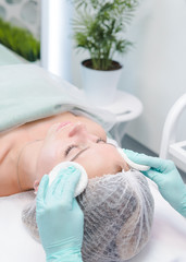 The female dermatologist cleaning face skin by cotton sponge to beautiful girl. Cosmetologist removing facial mask from female face