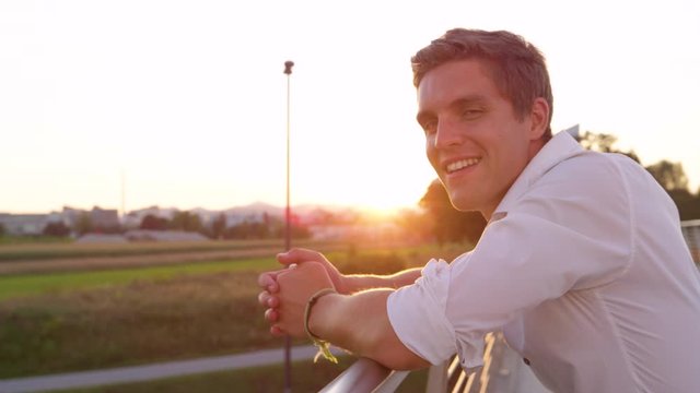 SLOW MOTION, LENS FLARE, CLOSE UP, DOF: Young businessman unwinding by looking around scenic sunlit nature smiles for the camera. Scenic shot of a summer sunrise shining on the confident male student