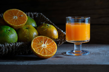 Oranges juice in glass and fresh orange in the brown basketry tray on the plank  which has dim light