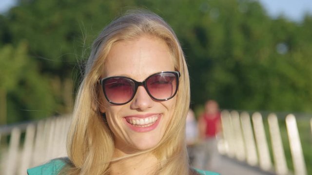 SLOW MOTION, CLOSE UP, PORTRAIT, DOF: Gorgeous blonde haired woman wearing sunglasses smiles while looking into the camera. Strikingly beautiful college student smiles during a fun day at the park