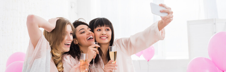 Obraz na płótnie Canvas panoramic shot of emotional multicultural girls with glasses of champagne taking selfie on smartphone during pajama party