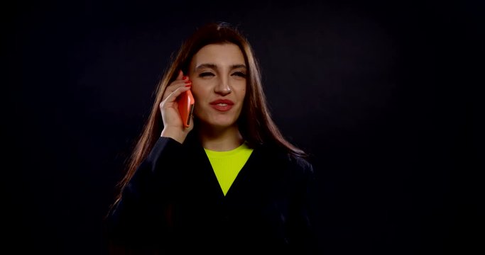 portrait of a brunette with bright lipstick on her lips, red nail Polish on her nails, and gold rings in her ears on a dark background. she talks on a smartphone in an orange case and laughs