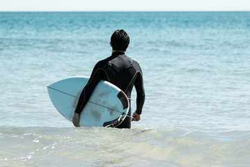 Back view of man carrying surfboard and walking into sea water. Young guy wearing wetsuit with seascape in background. Surfboarding and vacation concept.