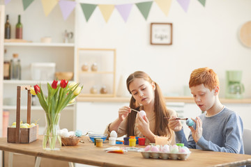 Two friends sitting at the table and painting eggs in colorful paints they preparing for Easter