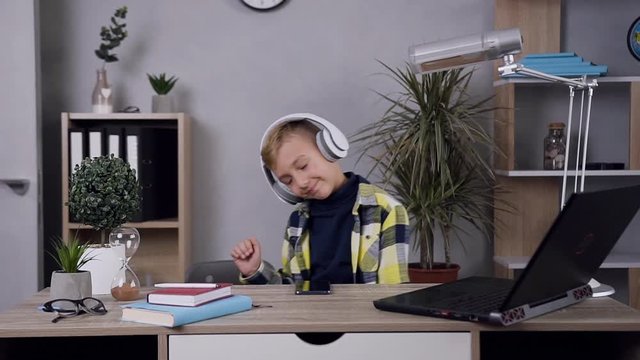 Portrait of attractive cheerful funny teen boy in headphones which enjoying music and dancing near the table