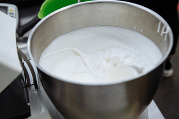 Whipped egg whites cream to perfect peaks in glass bowl with mixer and hand on blue wooden table. Step by step recipe of meringue cookies top view.