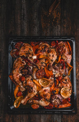 Roasted chicken legs with various vegetables and oranges on metal tray, wooden background, top view