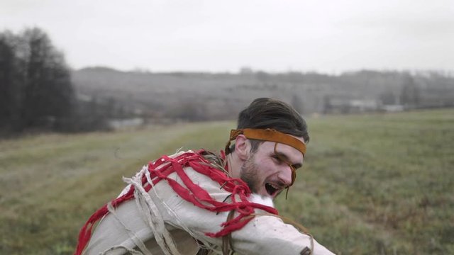 A man in a beige or white Native American suit is dancing and spinning, clothes are developing in the wind, a gray field, cloudy weather, against a forest