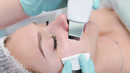 Fototapeta premium The female cosmetologist makes a procedure of ultrasonic cleaning face, close-up. Young woman in a beauty salon.