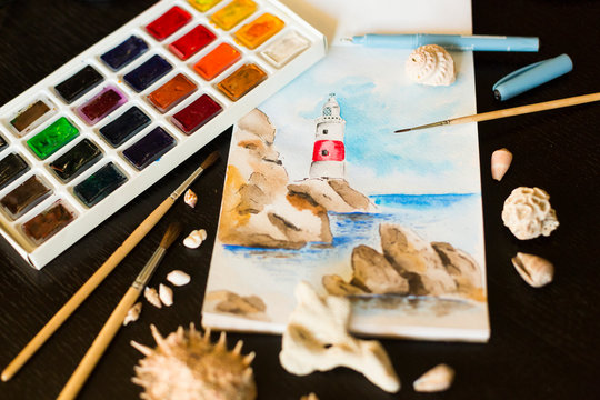 Colorful watercolor painting of old  lighthouse on cliff in the sea or ocean with palette, brushes, seashells and stones