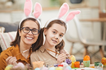 Portrait of happy mother and daughter in rabbit ears smiling at camera while sitting at the table with paints