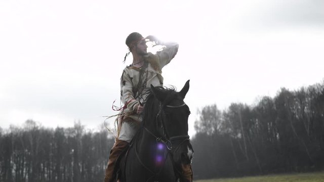  A man stands on a black horse in a field against a forest in the fall in cloudy and windy weather, clothes develop in the wind, Native American peers into the distance with a palm against his eyes