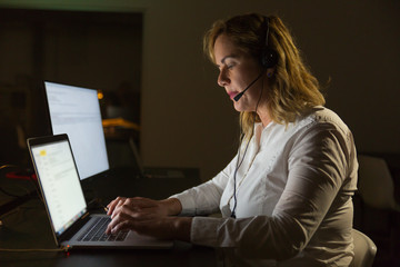 Call center operator working late in dark office. Side view of focused middle aged businesswoman using laptop computer in dark office. Client support concept