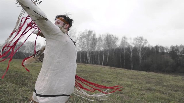 A man in a beige or white Native American suit is dancing and spinning, clothes are developing in the wind, a gray field, cloudy weather, against a forest