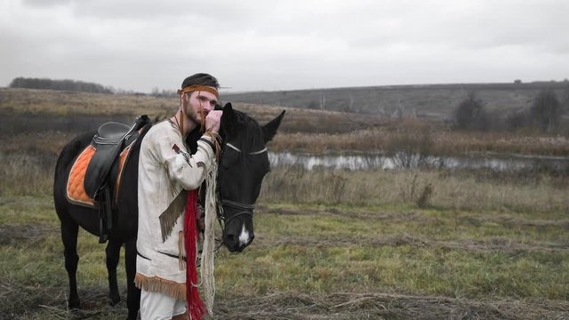 A young man in a beige suit with a bandage on his head. The Indian leaned his head against the black horse and stands, meaningfully thinks and strokes the horse in the face