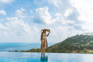 A beautiful girl in a swimsuit, a hat and a robe developing in the wind stands in a villa with a pool and beautiful views of the jungle and the sea. Thailand, Samui