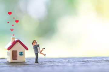 miniature couple and miniature house with red roof on wooden mock up over blurred green garden on...