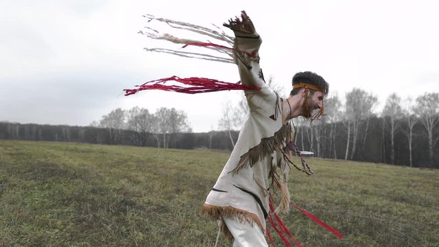 A man in a beige or white Native American suit is dancing and spinning, clothes are developing in the wind, a gray field, cloudy weather, against a forest