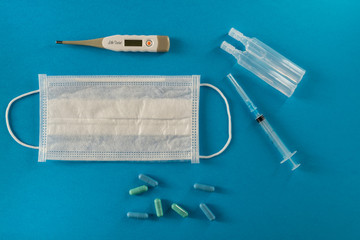White medical mask, thermometer, syringe, tablets and vials with medicine on the blue surface.