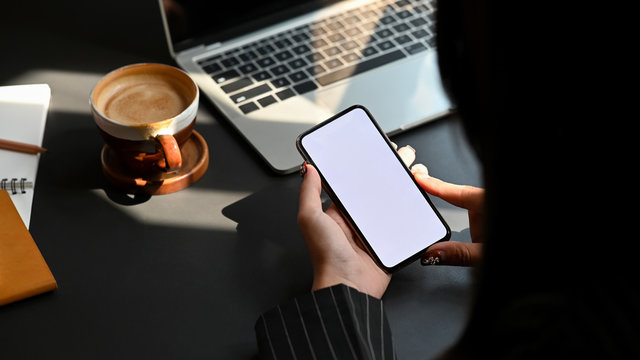Side shot of young businesswoman holding white blank screen smartphone in her hand while sitting at the modern working desk with laptop, notebook and coffee cup as background.