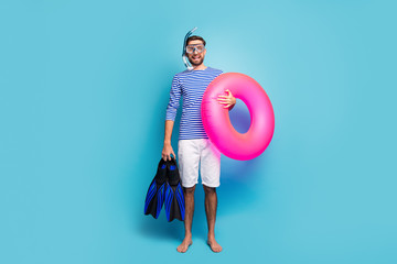 Full body photo of funny excited guy tourist swimmer hold underwater mask breathing tube flippers pink lifebuoy wear striped sailor shirt shorts isolated blue color background - Powered by Adobe