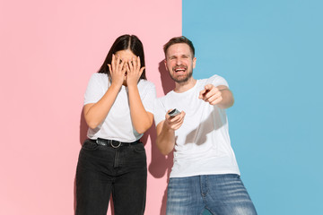 Who's in control. He chooses TV channel, she's angry. Young man, woman in casual on pink, blue bicolored background. Concept of human emotions, facial expession, relations, ad. Beautiful couple.