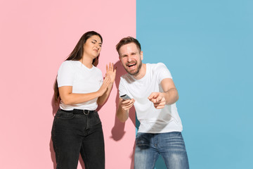 Who's in control. He chooses TV channel, she's angry. Young man, woman in casual on pink, blue bicolored background. Concept of human emotions, facial expession, relations, ad. Beautiful couple.