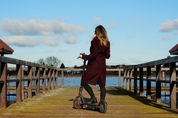 an unrecognizable woman with long hair in an elegant coat came by electric scooter on a wooden bridge over the lake and admires the landscape, copyspace