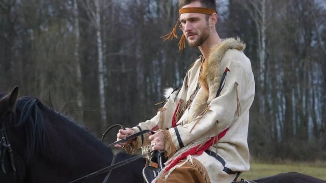 Young man in white beige Native American costume with red trousers and fur on his shoulder rides beautiful majestic black horse pulls the reins and gives a command against the background of the forest