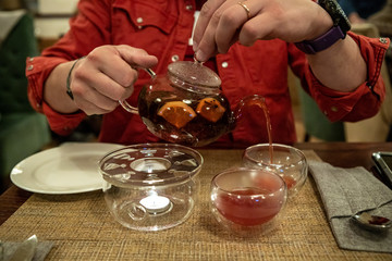A man in a red shirt pours fruit tea from a transparent teapot into cups in a cafe