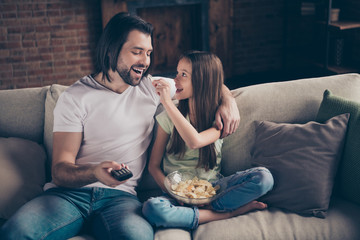 Photo of little funny lady giving handsome daddy eat chips sit cosy sofa good mood remote controller watch favorite film carefree teasing near mouth house room indoors