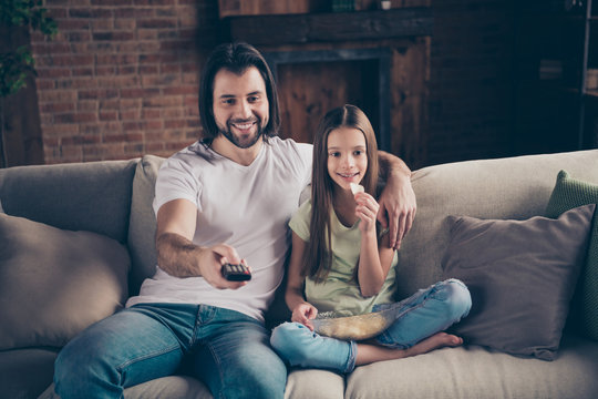 Profile photo of little funny lady handsome daddy sit cosy sofa good mood eating potato chips remote controller watch favorite film make volume bigger house room indoors
