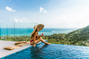 A beautiful girl in a red bathing suit is relaxing in a luxury villa by the pool with wine and in a hat taking cover from the sun. View of palm trees and the sea. Thailand, Koh Samui