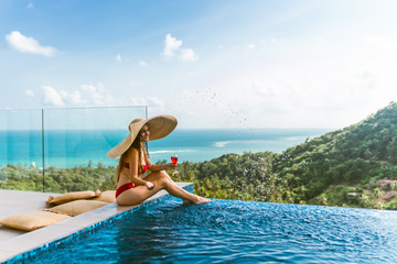 A beautiful girl in a red bathing suit is relaxing in a luxury villa by the pool with wine and in a hat taking cover from the sun. View of palm trees and the sea. Thailand, Koh Samui