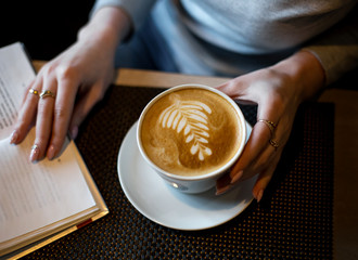 A white Cup of coffee in a woman's hands and an open book on the table