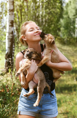 A beautiful happy blonde girl in a black t shirt and short blue shorts holds two puppies in her hands and kisses a puppy on a Sunny summer day