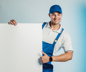 Mockup of smiling young worker man wearing blue uniform. Holding white empty banner in his hand and showing thumb up. Movement cool. Isolated on grey background with copy space. Business concept.