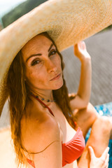 A beautiful girl in a red bathing suit is relaxing in a luxury villa by the pool with wine and in a hat taking cover from the sun. View of palm trees and the sea. Thailand, Koh Samui