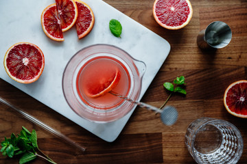 Mixing of refreshing cold summer cocktail with blood orange in a glass pitcher on a marble tray. Top view, flat lay composition.