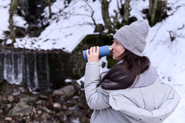 A young woman in winter clothes and a hat drinks hot tea from a thermos in the winter in the mountains. The concept of tourism and active lifestyle. Copy space.