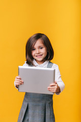 A caucasian girl in a gray dress and white shirt uses and plays with a tablet on a yellow background