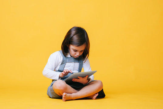 A caucasian girl uses and plays with a tablet. She is wearing a gray dress and white shirt on a yellow background